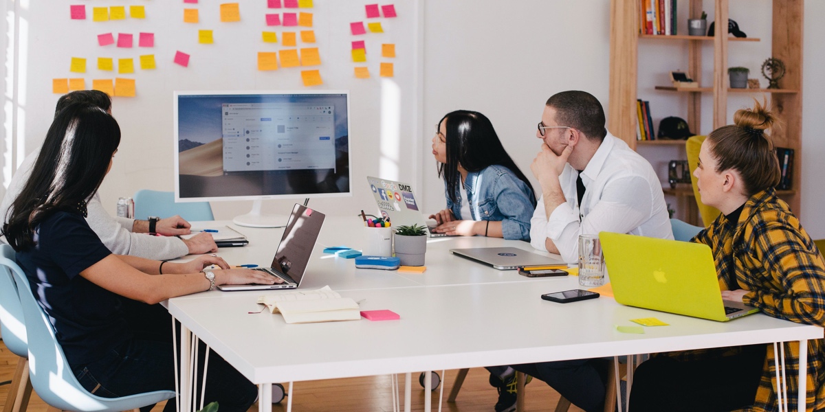 What's it like to be a UX researcher? Group of 5 designers gathered at a table, looking at a screen near a wall covered in sticky notes