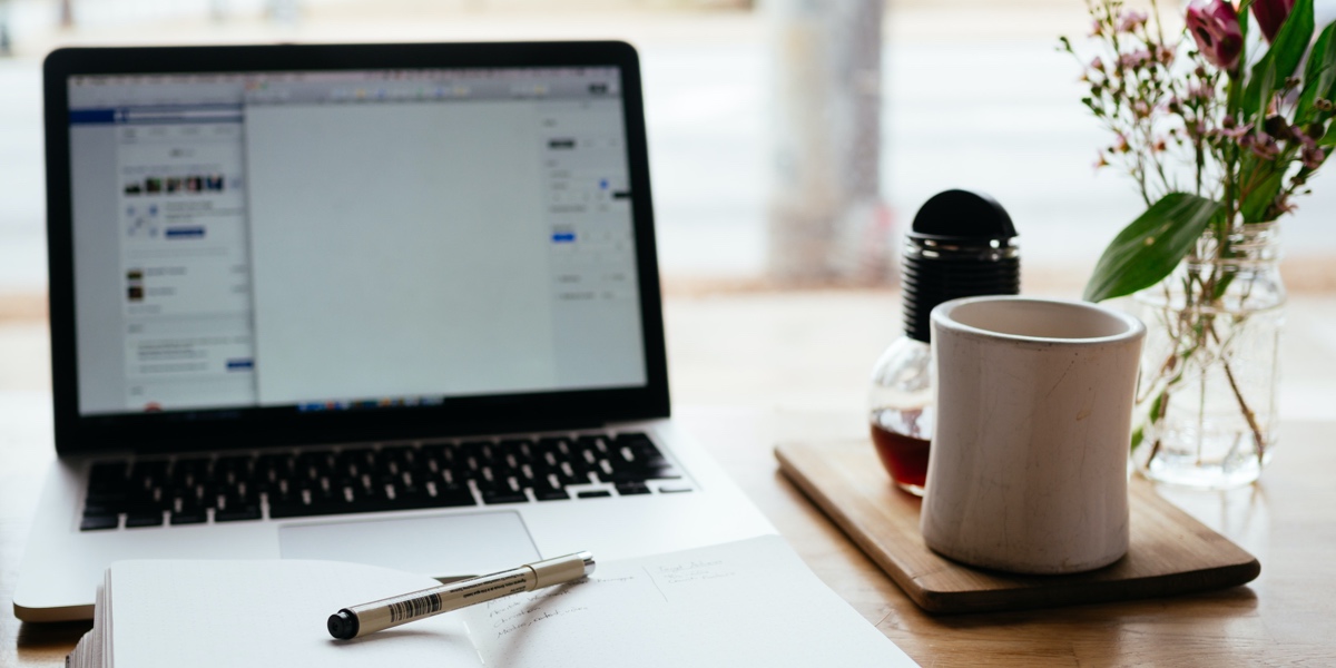 A desk area in a sunny room with a laptop, notebook, coffee cup, and vase of flowers